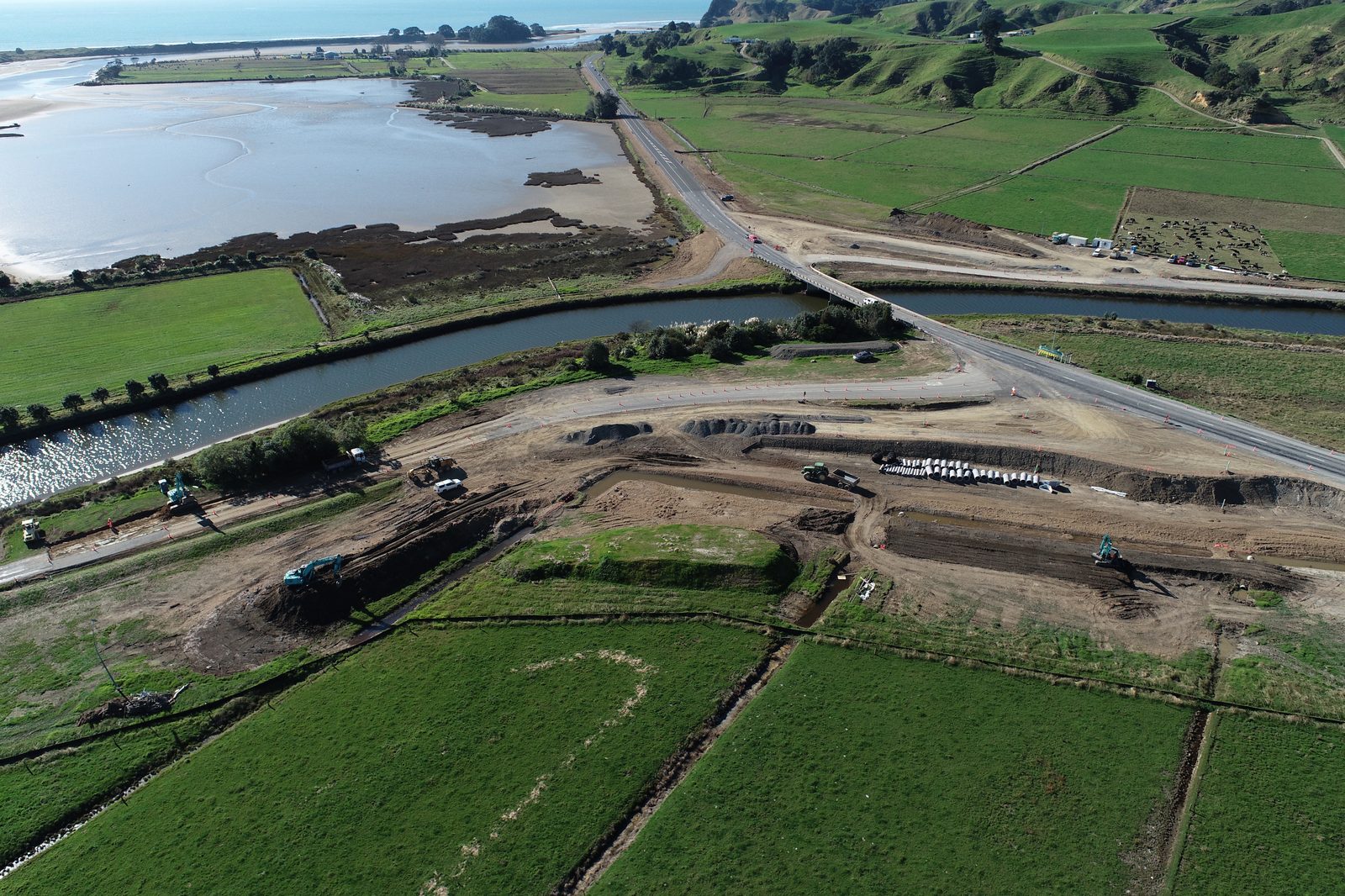 Aerial view of the SH2 and Ohiwa Beach Road teardrop intersection under construction — excavators shaping the earthworks alongside the estuary, eastern Bay of Plenty.
